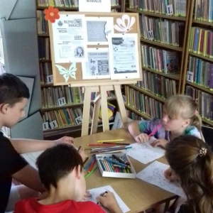 Dzieci kolorują kolorowanki. W tle tablica informacyjna i regały z książkami.