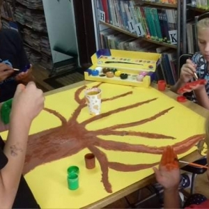 Dzieci malują drzewo i dłonie, w tle regały z książkami.