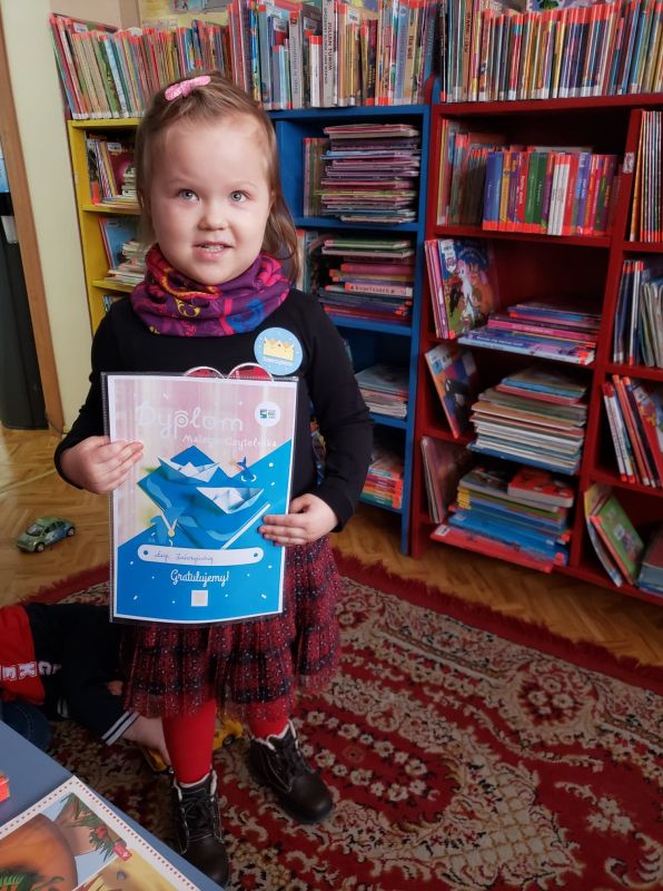 Mała dziewczynka znajduje się w bibliotece, stoi na tle kolorowych książek  trzymając w ręku dyplom otrzymany w ramach projektu Mała książka wielki człowiek.